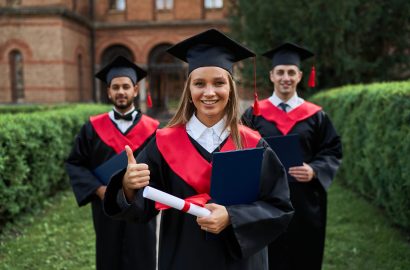 Graduate female shows like with her friends in graduation gowns holding diploma and smiling at camera.
